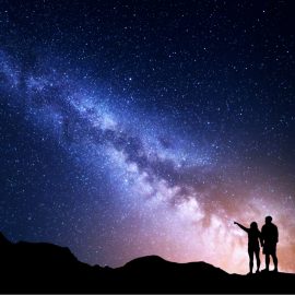 Two travelers stand on the top of a mountain, pointing at the Milky Way