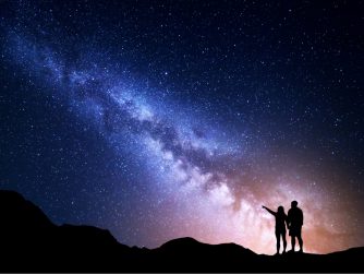 Two travelers stand on the top of a mountain, pointing at the Milky Way