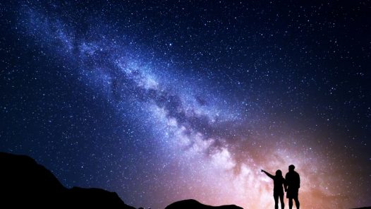 Two travelers stand on the top of a mountain, pointing at the Milky Way