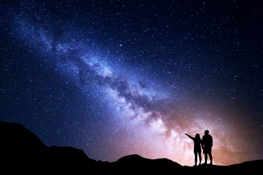 Two travelers stand on the top of a mountain, pointing at the Milky Way